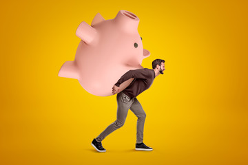 Young man in casual clothes carrying a big pink piggy bank on his back on yellow background