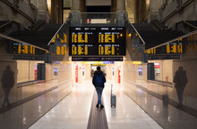 Passenger With A Backpack And A Suitcase Beside Her Is Standing Below A Timetable Board In A Giant Hall Of A Railway Station In Milan, Italy. Text: "Arrivals, Departures". Solo Traveling Concept.