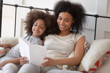 Wall Mural - Happy african mother reading book relaxing in bed with daughter