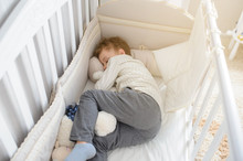 Top View Of A Blond Seven Years Boy Sleeping In His Sister White Wooden Cot In White Shirt And Grey Trousers With Toys In His Arms