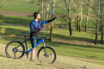 Cyclist in shorts and jersey on a modern carbon hardtail bike with an air suspension fork rides off-road on green hills near the forest	