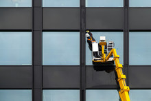 Male Window Cleaner Cleaning Glass Windows On Modern Building High In The Air On A Lift Platform. Worker Polishing Glass High In The Air