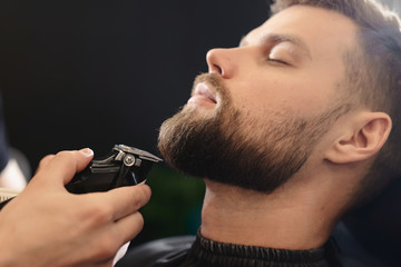 Getting perfect shape. Close-up side view of young bearded man getting beard haircut by hairdresser or barber at barbershop