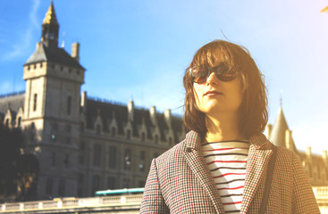 Portrait of young beautiful girl in sunglasses standing on the embankment of river Seine with the palace on the background in the center of Paris, France. Summer vacation tour in Europe. Stylish woman