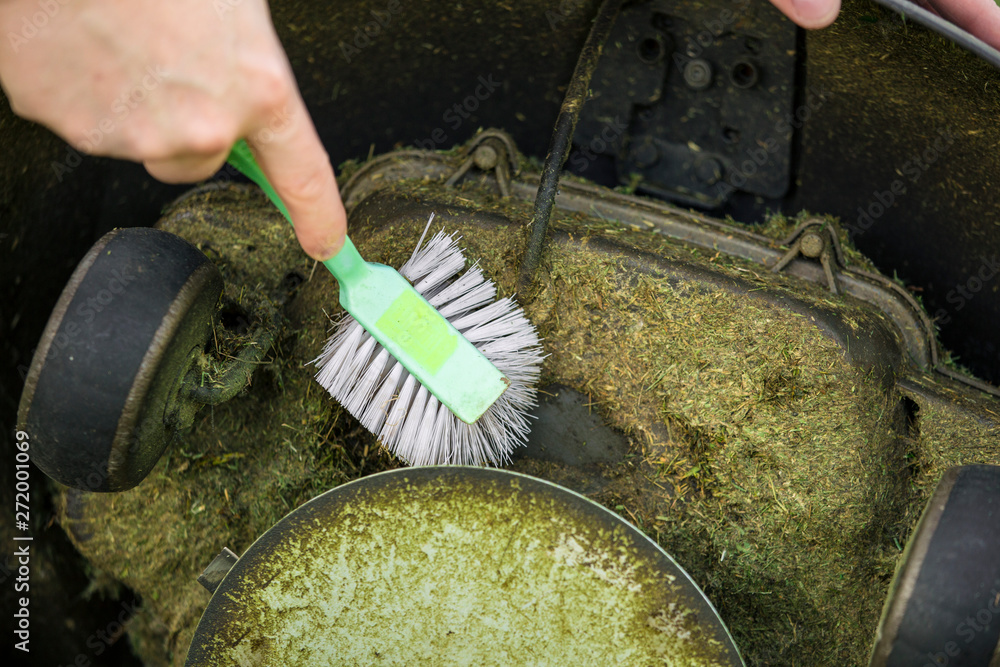 Close up view of man hand brushing off layer of wet grass stuck under automated lawnmower, maintenance concept.