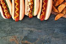 Hot Dogs With Toppings And Potato Wedges. Top Border, Overhead View On A Dark Stone Background With Copy Space.
