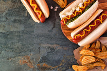 Hot Dogs With Toppings And Potato Wedges. Corner Border, Above View On A Dark Stone Background With Copy Space.