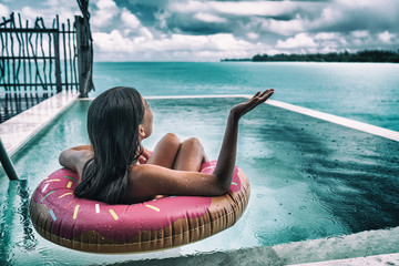 Sad woman on rainy day vacation bad holiday rain on travel vacations in Caribbean. Upset woman in donut float on swimming pool.