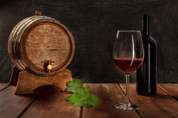 Canvas Print - A glass of red wine with a bottle, a wine barrel, and vine leaves, side view on a dark rustic background, low key photo with a place for text