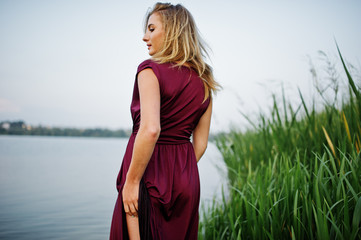 Wall Mural - Blonde sensual woman in red marsala dress standing in water of lake with reeds and show her nude leg.