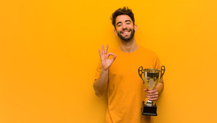 Wall Mural - Young man holding a trophy cheerful and confident doing ok gesture