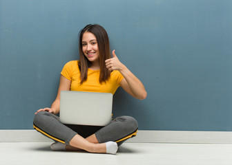 Wall Mural - Young woman sitting on the floor with a laptop smiling and raising thumb up