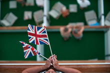 A Lady Holds Two Union Jack Flags. Viewed From Above With Cruise Ship Outside Balconies Blurred Below
