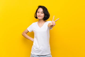 Asian young woman over isolated yellow wall smiling and showing victory sign