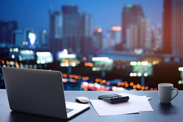 Laptop on table in office room on window city background.Computer and white paper,calculator with a coffee cup on a black desk