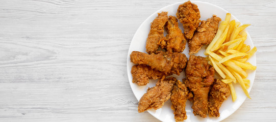 Wall Mural - Tasty fried chicken legs, spicy wings, French fries and chicken strips on a white plate over white wooden surface, top view. Flat lay, overhead, from above. Copy space.