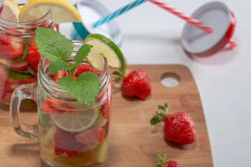 Two mugs of fruit water with strawberries mint and lemon stand on a wooden stand next to fresh strawberries and mugs from mugs, white background.