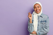 Carefree optimistic female with toothy smile, being in high spirit, wrapped in white scarf, wears fashionable denim jacket, looks away, poses over purple background, blank space area for information