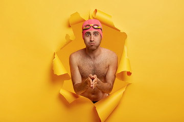 Topeless young male blows cheeks, keeps palms pressed together catches breath, prepares for diving under water, wears special goggles and swimhat, stands in yellow wall hole with ragged edges