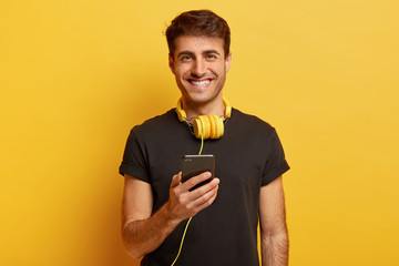 Portrait of happy Caucasian man enjoys fantastic sound and quality of headphones, holds modern cell phone, listens music, smiles joyfully, isolated over yellow background. People and pastime concept