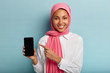 © wayhome.studio  - Muslim lady holds smartphone, shows blank screen to insert text or your information, wears pink hijab and white shirt, advertises new gadget, smiles happily, isolated over blue wall. Selective focus