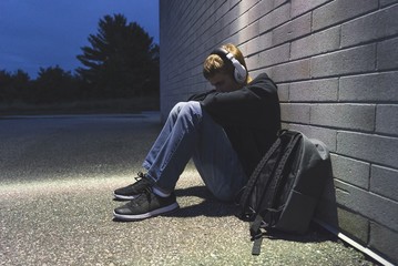 Wall Mural - Sad teenage boy sitting on the ground against a brick wall at night. He is listening to music through a pair of headphones.