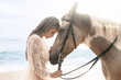 Happy fashionable young woman in a white dress posing with a horse on the beach.