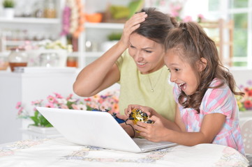 Wall Mural - Portrait of cute little girl with young mother playing computer game with laptop