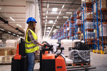 Woman Warehouse Worker Operating Forklift Machine In Large Distribution Warehouse Center. Manual Worker Relocating Goods In Factory Storage Area.