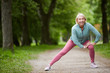 © Seventyfour - Mature cheerful blonde female in activewear stretching legs while doing physical exercises on road in city park among green trees
