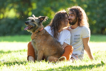 Wall Mural - Happy beautiful curly young couple sits on the grass with with  a cute dog