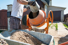Builder Working With Shovel During Concrete Cement Solution Mortar Preparation. Construction Worker With A Bucket In His Hands Loads A Concrete Mixer.orange Concrete Mixer Prepares Cement Mortar