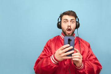 Wall Mural - Surprised man with a beard in the headphones, holds a smartphone in his hands and looks at the camera with a shocking face. Emotional man listens to music and uses a smartphone on a blue background