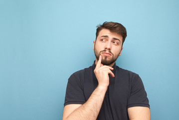 Wall Mural - Man with a beard and dressed in a dark T-shirt thinks on a blue background and looks sideways on сopyspace. Thoughtful adult man is isolated on a blue background.