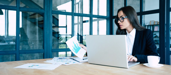 Successful confident beautiful young brunette businesswoman sitting at modern workplace and working with business papers and graphics