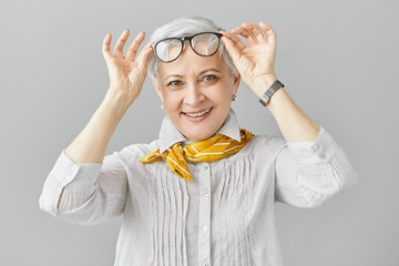 Beautiful fashionable Caucasian woman pensioner with farsightedness taking off her eyeglasses to focus on closer objects, smiling broadly at camera. Mature people, aging and vision problem concept