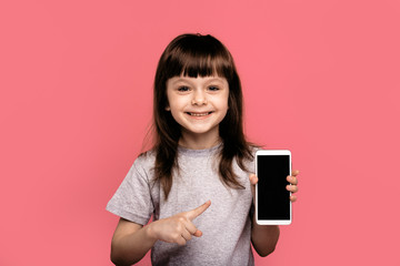 Look at this cell phone! Pleased happy little girl showing blank screen of smart sell phone, modern device, dressed in fashionable t-shirt, isolated over pink background. Technology concept