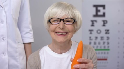 Wall Mural - Oculist proposing carrot to smiling elderly lady, natural vitamins for sight