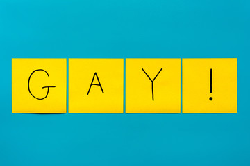 Handwritten black inscription gay with exclamation mark on yellow square stickers on a blue background close-up, horizontal orientation.