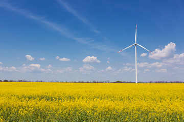 in the yellow canola field stands a white turbogenerator, against the sky with clouds, the concept