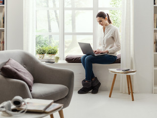 Wall Mural - Young woman sitting next to a window and connecting with her laptop