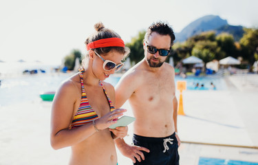 A couple in swimsuit by the swimming pool on summer holiday, using smartphone.