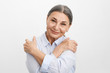 © shurkin_son - Horizontal shot of charming relaxed cheerful elderly senior female in blue shirt keeping hands on shoulders with arms on chest, hugging herself, looking at camera with joyful peaceful smile