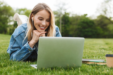 Wall Mural - Photo of gorgeous caucasian woman smiling and using laptop with earphones while lying on green grass in park