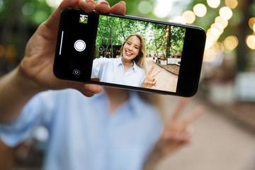 Canvas Print - Photo of cute smiling woman taking selfie photo on smartphone gesturing peace sing while walking in park