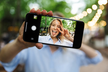 Wall Mural - Photo of blonde nice woman selfie photo on smartphone with sticking out her tongue and gesturing peace sing