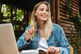 Portrait of blonde smiling woman writing in diary while using laptop in cafe outdoors