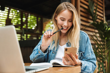 Wall Mural - Portrait of caucasian smiling woman writing in diary while using laptop and cellphone in cafe outdoors