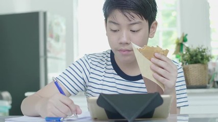 Wall Mural - Asian boy Sits on a chair at home doing homework using a tablet to find answers While in the hand holding the food