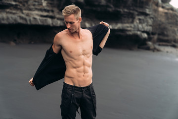 Wall Mural - Sexy fitness male model in black pants and shirtless posing on black sandy beach.